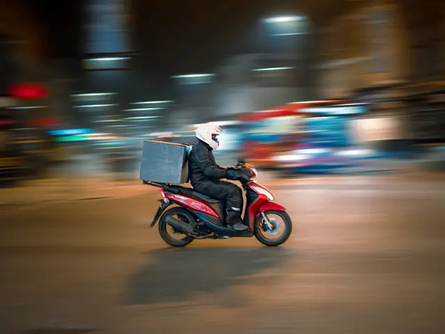 Entregador de scooter com o baú de entrega, à noite, numa rua movimentada.
